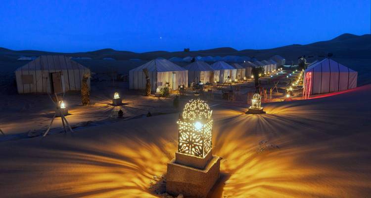 Fila de tiendas de lujo del desierto suavemente iluminadas bajo un cielo crepuscular estrellado en medio de dunas de arena ondulantes.