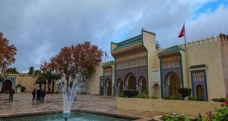 Puerta decorativa de palacio con arcos de azulejos coloridos, banderas marroquíes y una fuente en primer plano en un día nublado.