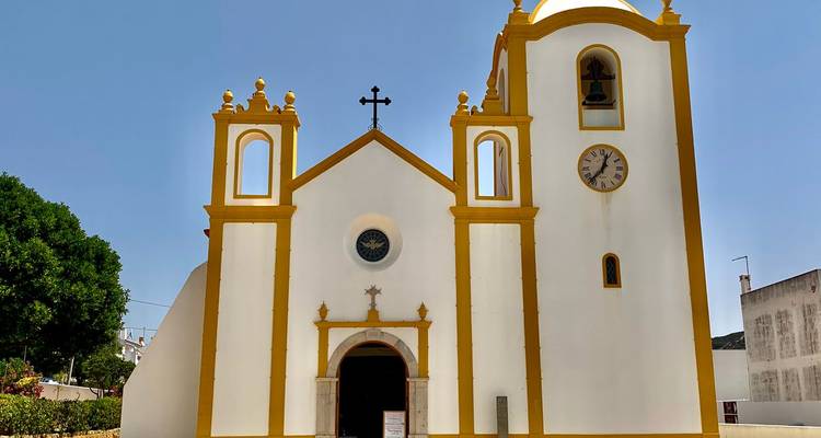 Strahlend weiße Kirche mit gelben Verzierungen, Zwillingstürmen und Uhr vor einem klaren blauen Himmel