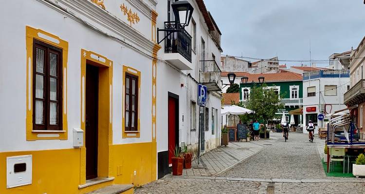 Bunte portugiesische Dorfstraße mit weiß getünchten und gelb verzierten Gebäuden, Kopfsteinpflaster und einigen Fußgängern und Radfahrern.