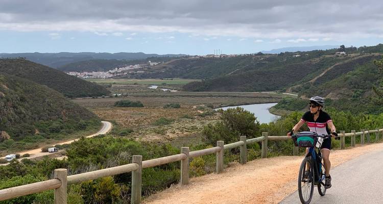 Ein Radfahrer fährt eine malerische Bergstraße entlang mit Blick auf ein gewundenes Flusstal mit bewaldeten Hügeln und einem entfernten Dorf.