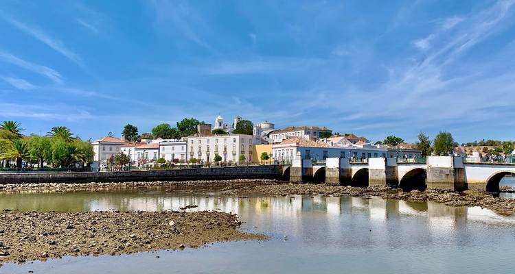 Historische Flussstadt mit einer mehrbogigen Steinbrücke, die sich in ruhigem Gezeitenwasser unter blauem Himmel spiegelt.