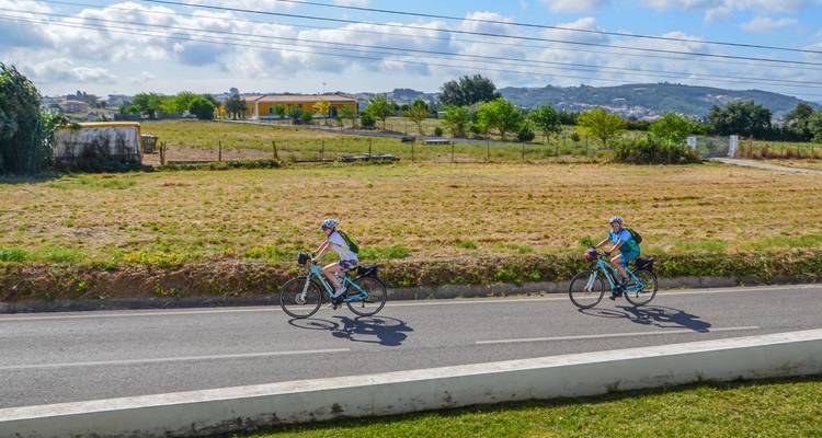 Zwei Rennradfahrer fahren eine Landstraße entlang und passieren abgeerntete Felder und entfernte Hügel.