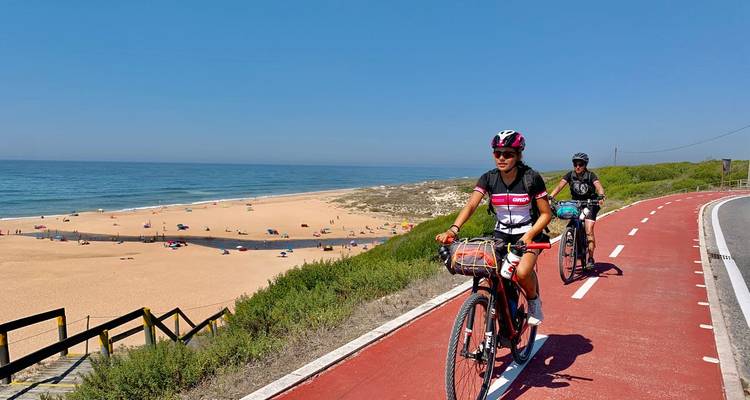 Radfahrer fahren auf einem roten Küsten-Radweg neben einem sandigen Strand und blauen Atlantikgewässern.