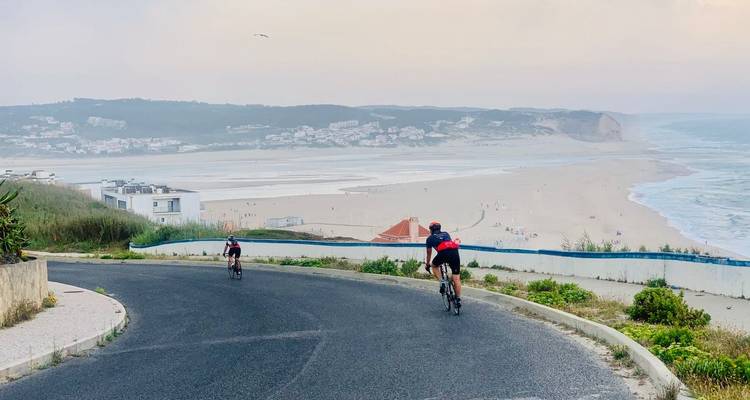 Zwei Radfahrer fahren eine Küstenstraße hinunter, die einen breiten Sandstrand und eine Küstenstadt überblickt.