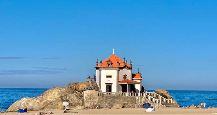 Kleine Kapelle am Meer mit orangefarbenem Dach, die auf Felsen thront, vor einem klaren blauen Himmel.