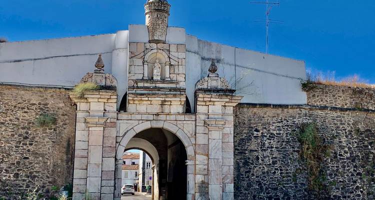 Puerta de piedra con escudo decorativo que conduce a una ciudad fortificada, contra un cielo azul profundo.