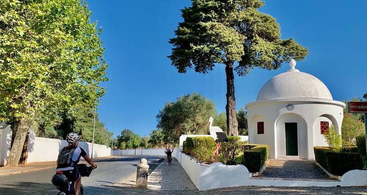 Los ciclistas pasan junto a una pequeña capilla blanca de cúpula entre setos cuidados y árboles altos en un día soleado.