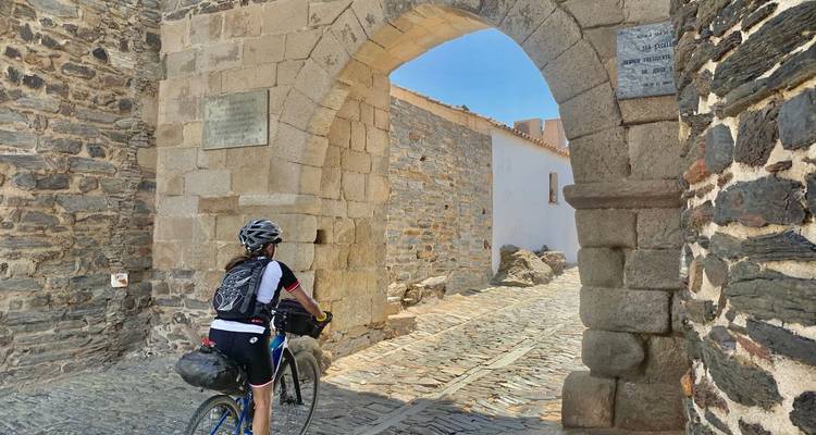 Radfahrer schiebt Fahrrad durch einen Steinbogen eines alten Dorftors an einem sonnigen Tag.