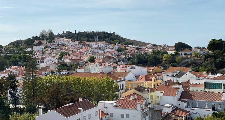 Panoramablick vom Hügel auf eine portugiesische Stadt mit dicht gedrängten Terrakottadächern, einer Kirche auf einer Anhöhe und belaubten Bäumen darunter.