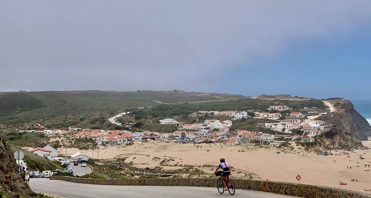 Radfahrer, der eine gewundene Küstenstraße hinab zu einem Sandstranddorf fährt, das von zerklüfteten Klippen und rollender Brandung umgeben ist.