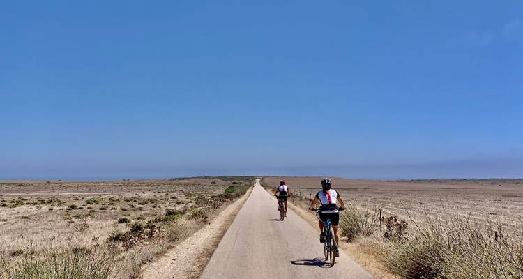 Zwei Radfahrer fahren auf einem schmalen asphaltierten Weg durch eine weite, trockene Ebene unter einem wolkenlosen blauen Himmel.