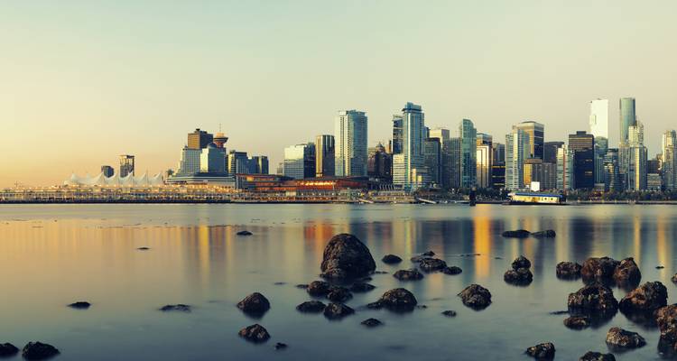 Ruhiges Wasser spiegelt eine moderne Skyline bei Tagesanbruch mit felsigem Vordergrund wider.