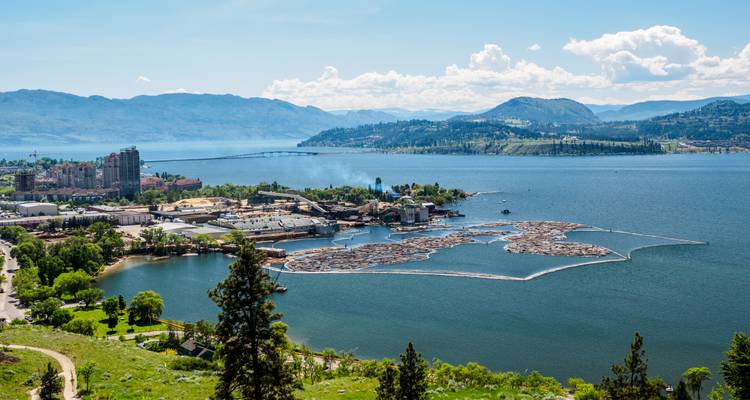 Seeuferstadt mit schwimmenden Baumstammflößen und Bergkulisse an einem sonnigen Tag.