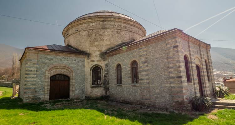 Église en pierre historique à dôme rond avec fenêtres en arc en ciel, située sur fond de montagnes et de ciel bleu.