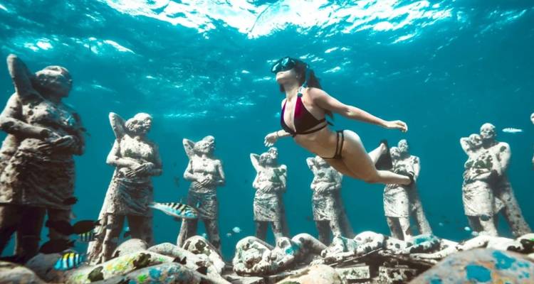 Un buceador con snorkel se desliza a través de un jardín de esculturas submarinas rodeado de vida marina en aguas azul cristalinas.