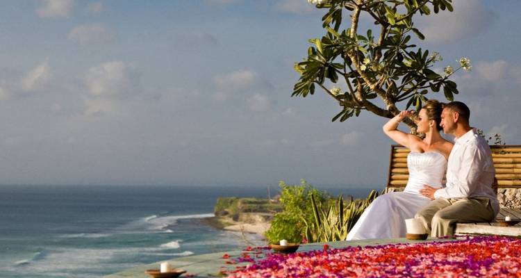Pareja en traje de boda se relaja en un banco en la cima de un acantilado con vista a un vasto panorama oceánico bajo cielos azules.