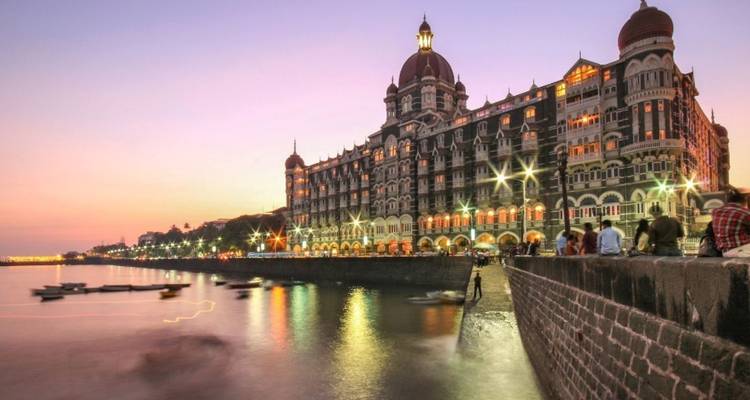 El iluminado Hotel Palacio Taj Mahal bordeando el puerto de Mumbai al atardecer pastel con reflejos borrosos de barcos en el agua.
