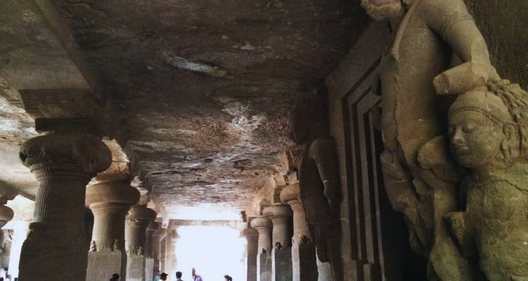 Sala de piedra tenuemente iluminada dentro de las Cuevas de Elephanta con enormes pilares tallados en roca y deidades esculpidas silueteadas contra una abertura.