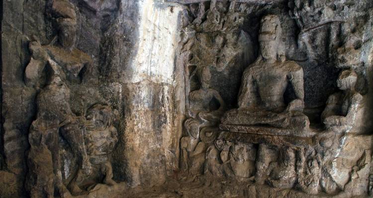Panel de bajorrelieve antiguo dentro de las Cuevas de Elephanta que muestra figuras de piedra sentadas y de pie rodeadas de roca erosionada.