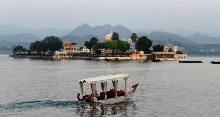 Barco sereno navegando por el lago Pichola hacia el palacio insular de Jag Mandir salpicado de árboles con las brumosas colinas Aravalli de fondo.