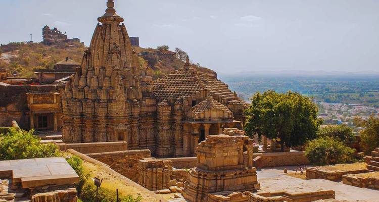 Templos de piedra rugosa y murallas del Fuerte de Chittorgarh contemplando una vasta llanura desértica bajo un cielo brumoso del mediodía.