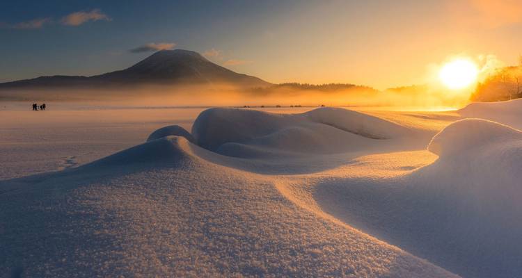 Golden sunrise lights fresh snow drifts on a frozen lake with a solitary cone-shaped volcano in the distance.