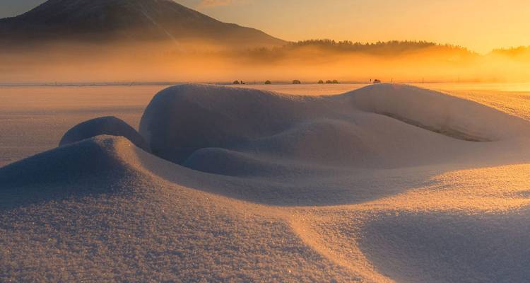 Gouden zonsopgangslicht streelt ongerepte sneeuwhoopen onder een verre berg.