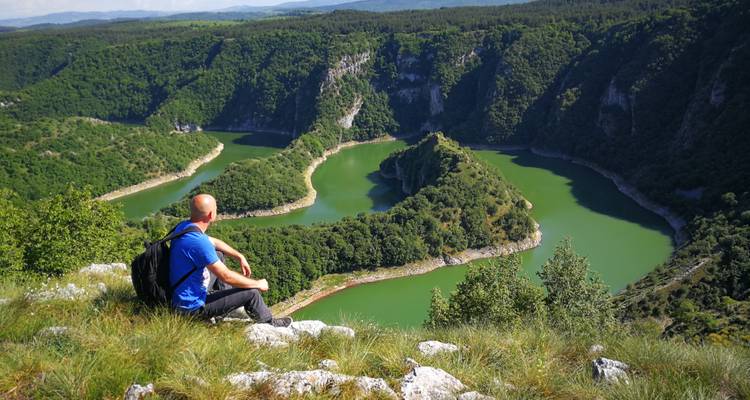 Randonneur assis au bord d'une falaise surplombant les méandres verts spectaculaires du canyon de la rivière Uvac au milieu de collines boisées vallonnées.
