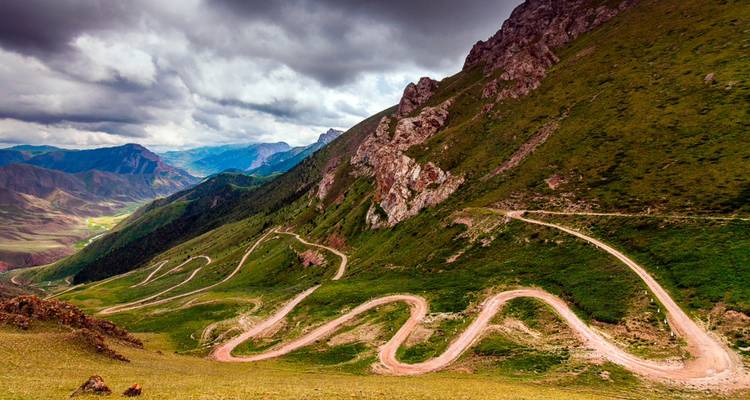 Une route de terre sinueuse serpente le long d'une montagne verte escarpée sous des nuages spectaculaires au Kirghizistan