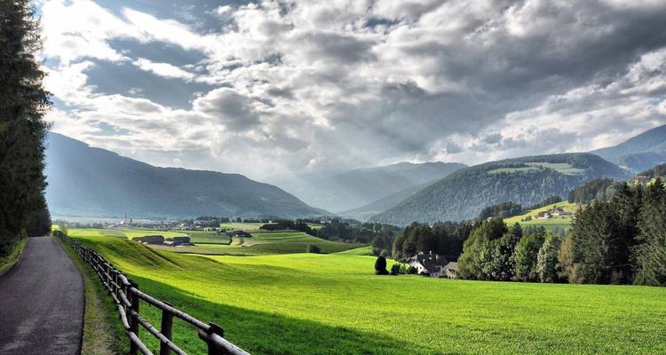 Glooiende groene weiden en lappendekenvelden strekken zich uit tussen mistige bergketens onder dramatische wolken.