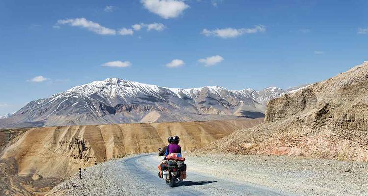 Motorradfahrer fahren auf einer Schotterbergstraße in Richtung schneebedeckter Gipfel unter einem strahlend blauen Ladakh-Himmel.