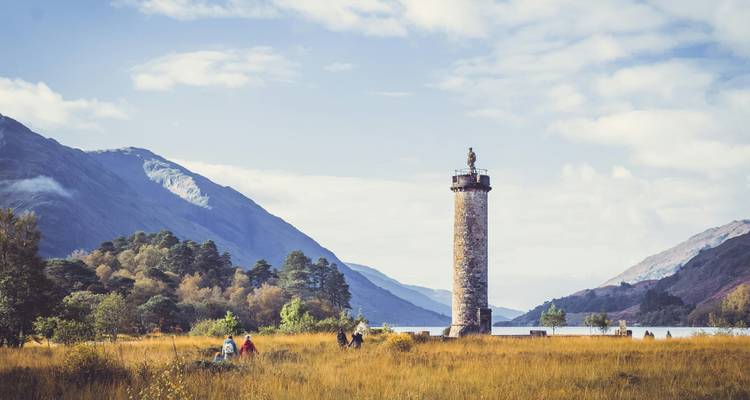Un grand monument de pierre s'élève d'une prairie dorée au bord d'un loch écossais tranquille avec des montagnes en arrière-plan.