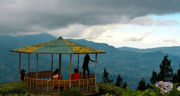 Menschen genießen die Aussicht von einem hölzernen Pavillon mit Blick auf sanfte blaue Berge unter dramatischen Wolken.