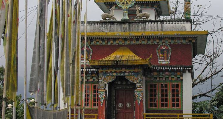 Farbenfroher buddhistischer Schrein auf einem Hügel mit kunstvollen Wandmalereien und flatternden Gebetsfahnen.