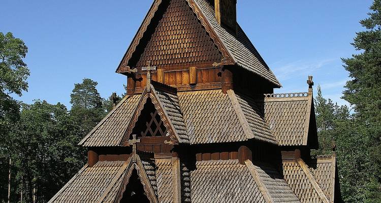 Église en bois debout norvégienne traditionnelle en bois sombre nichée parmi les arbres sous un ciel bleu.