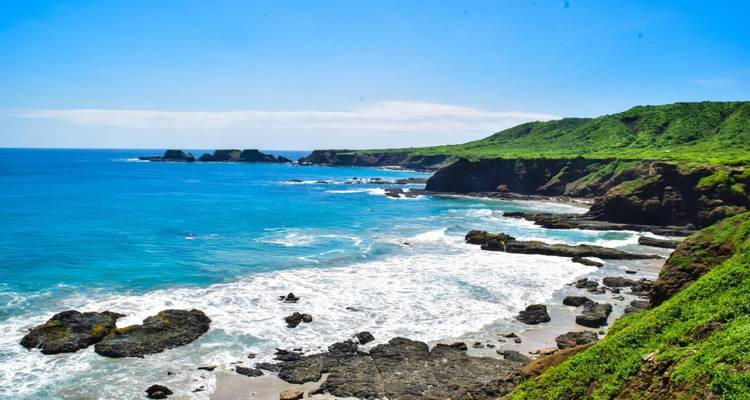 Vibrantes acantilados costeros verdes descienden hacia olas rompientes y calas rocosas bajo un cielo azul brillante.