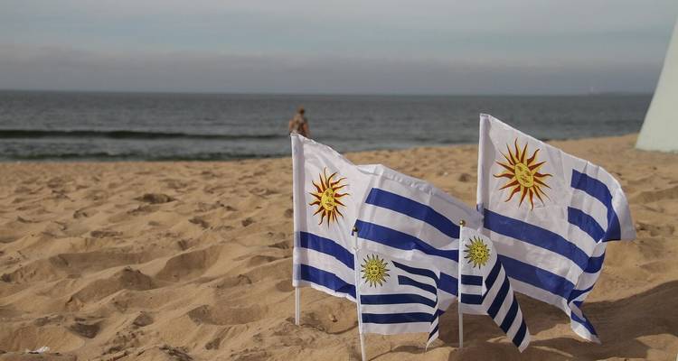 Kleine uruguayische Flaggen flattern im goldenen Strandsand mit ruhigen Meereswellen und einem einsamen Badegast dahinter.