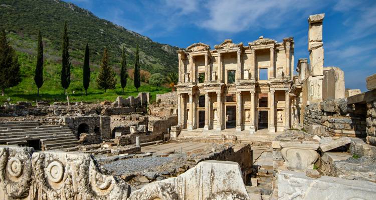 Oude marmeren zuilen van de ruïnes van de Bibliotheek van Celsus omlijst door groene heuvelflank en blauwe lucht.