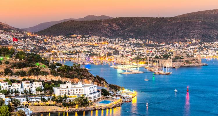 Zonsondergangzicht op de haven van Bodrum met kasteel, huizen op de heuvel en boten verlicht tegen turkooise wateren.