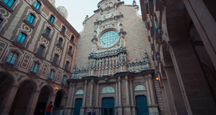 Façade de pierre et portail sculpté de la basilique de Montserrat s'élevant entre de hauts murs de monastère dans une cour étroite.