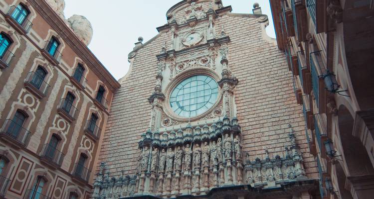 Vue en contre-plongée de la façade de pierre ornée de la basilique de Montserrat avec sa grande fenêtre circulaire et ses sculptures détaillées.