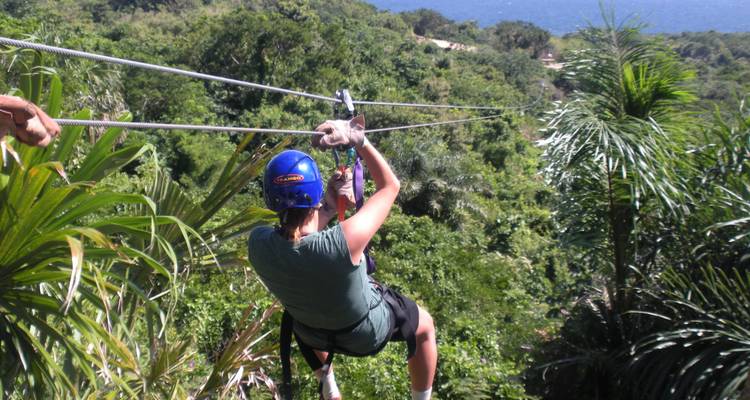 Traveler zip-lining over dense tropical forest with distant water view.