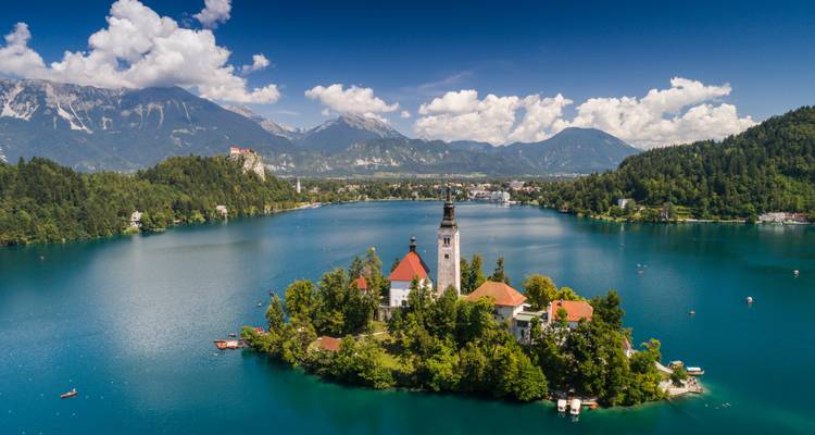 Panorama aérien du lac de Bled avec son île surmontée d'une église, les forêts environnantes, un château perché sur une falaise et des sommets alpins sous un ciel bleu profond.