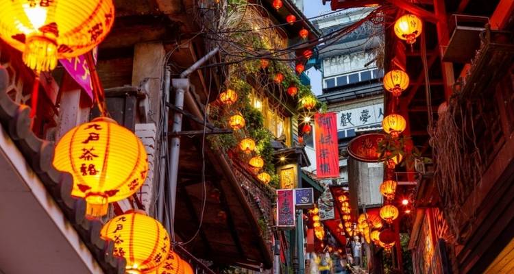 Rue étroite et ancienne de Jiufen illuminée par des rangées de lanternes rouges et des enseignes colorées de magasins au crépuscule.