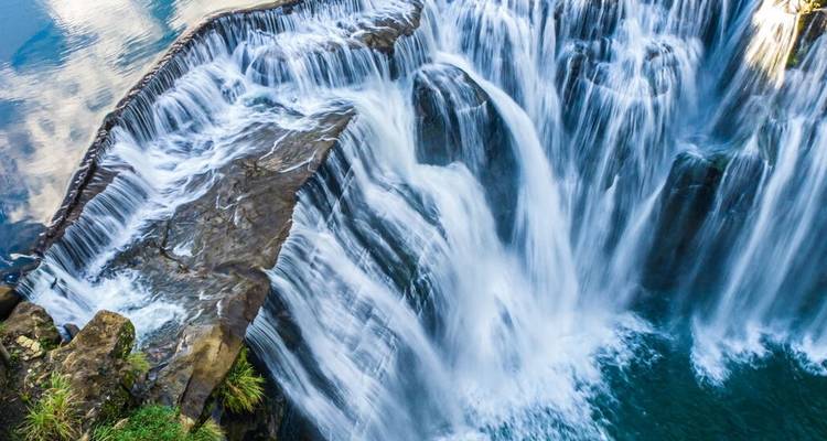 Puissant rideau d'eau qui plonge par-dessus les rebords rocheux dans un bassin turquoise sous une cascade forestière.