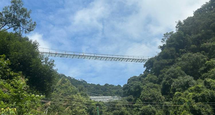 Een lange glazen hangbrug strekt zich uit tussen twee beboste heuvels onder een gevlekte blauwe hemel.