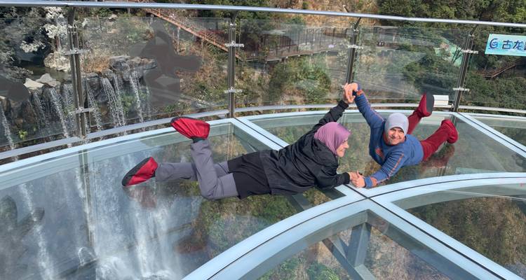 Twee bezoekers liggen speels op een transparant glazen platform hoog boven een waterval, met beschermende schoenovertrekken aan.