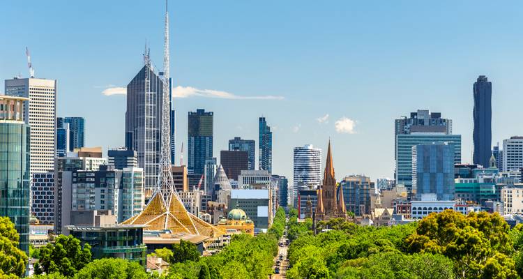 Horizonte de Melbourne enmarcado por frondosos árboles verdes con la aguja del Centro de las Artes prominente bajo cielos despejados