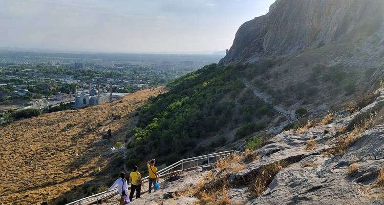Drei Wanderer steigen einen felsigen Pfad mit Panoramablick auf die Stadt und Moscheen in Osch hinab.